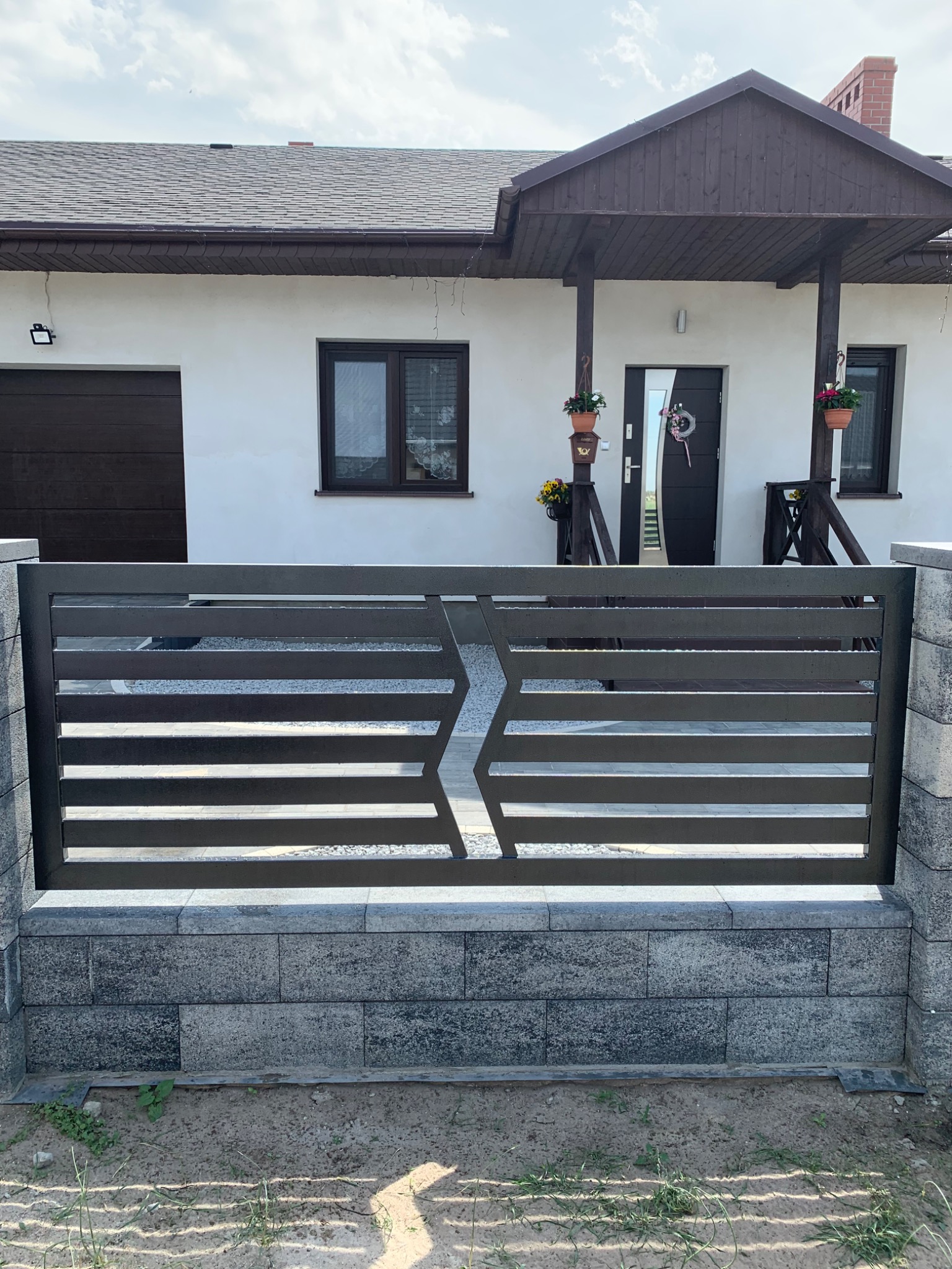 Nowoczesne, poziome przęsło ogrodzeniowe z metalu w kolorze grafitowym, zamontowane na murku z szarych bloczków betonowych, na tle białego domu z ciemnymi elementami wykończenia.