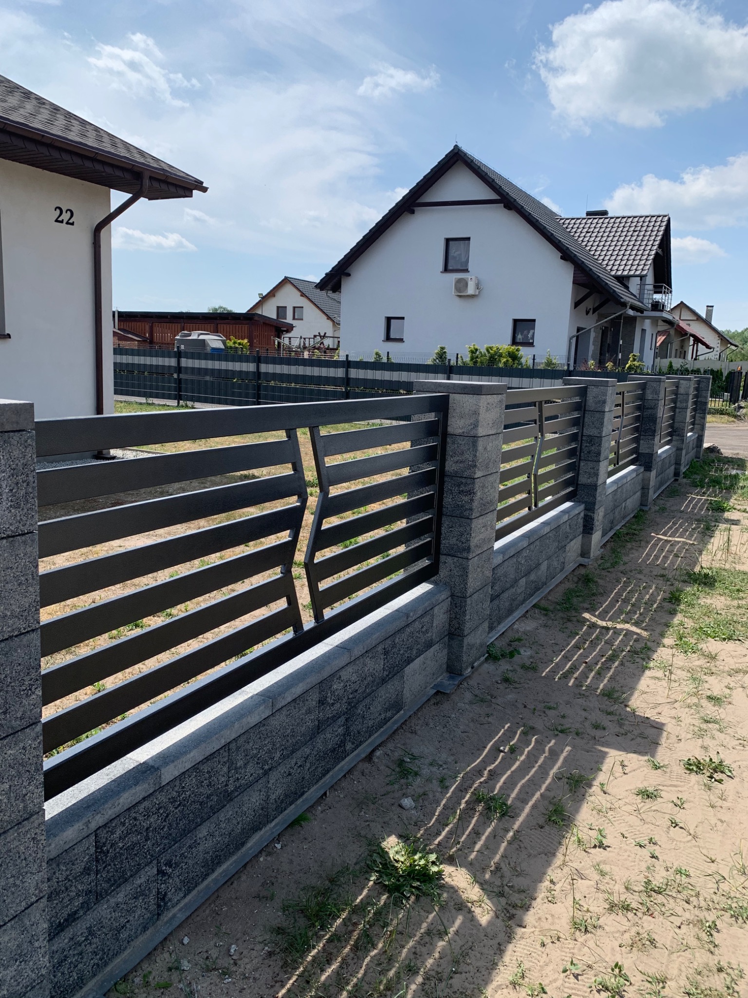 Nowoczesne ogrodzenie panelowe z poziomych, grafitowych elementów zamontowane na słupkach z szarej kostki łupanej, na tle domów jednorodzinnych w słoneczny dzień.
