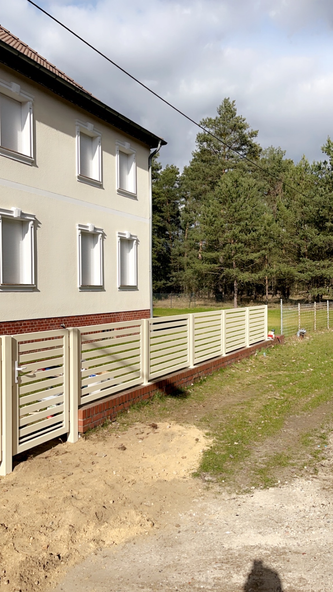 Nowoczesne, beżowe ogrodzenie panelowe z poziomymi szczeblami, zamontowane na niskiej, ceglanej podmurówce, przylegające do jasnego budynku mieszkalnego z widocznymi oknami i drzewami w tle.