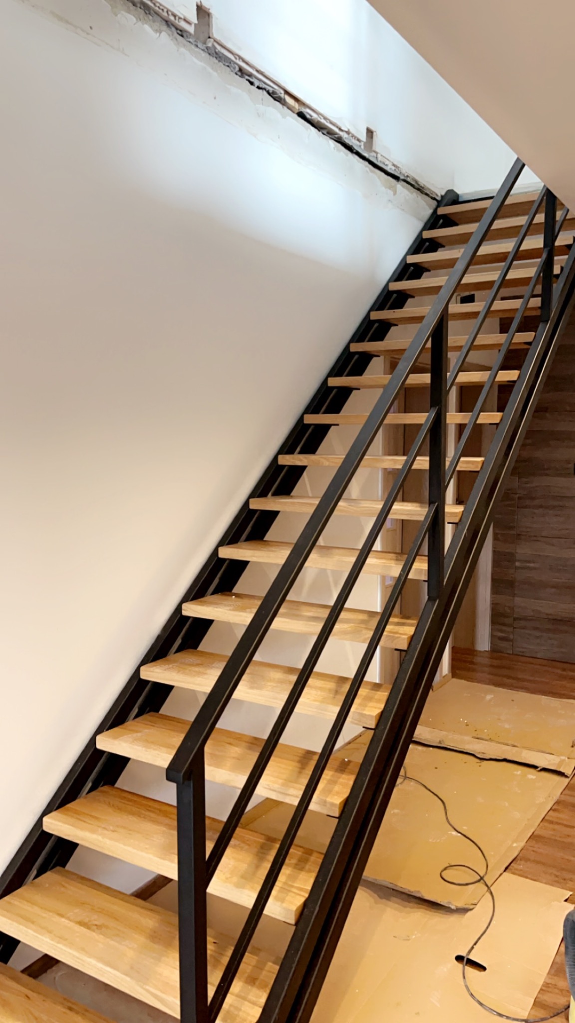Wnętrze z drewnianymi schodami o metalowej konstrukcji w trakcie montażu, widoczne zabezpieczenia podłogi i niedokończone wykończenie ściany.