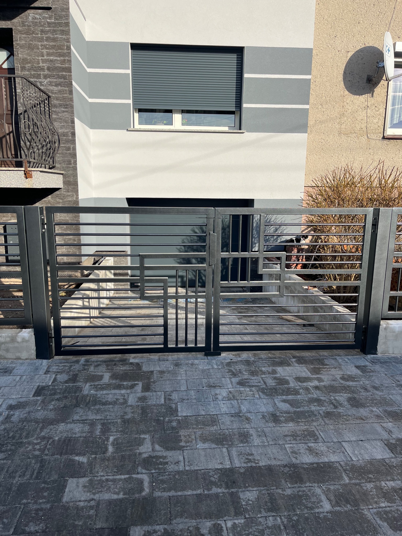 Nowoczesna, pozioma brama wjazdowa z metalowych profili w kolorze antracytowym, z geometrycznym wzorem, na tle dwukondygnacyjnego budynku z szarą elewacją i kostki brukowej.