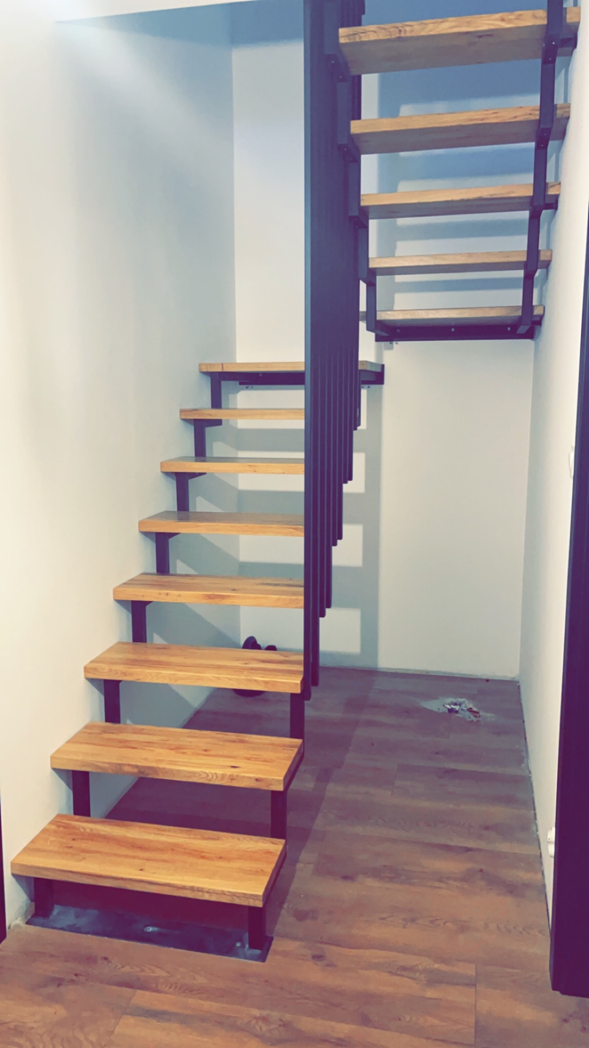 Nowoczesne schody z drewnianymi stopniami i czarną, metalową konstrukcją nośną, zamontowane w jasnym pomieszczeniu z panelami podłogowymi.