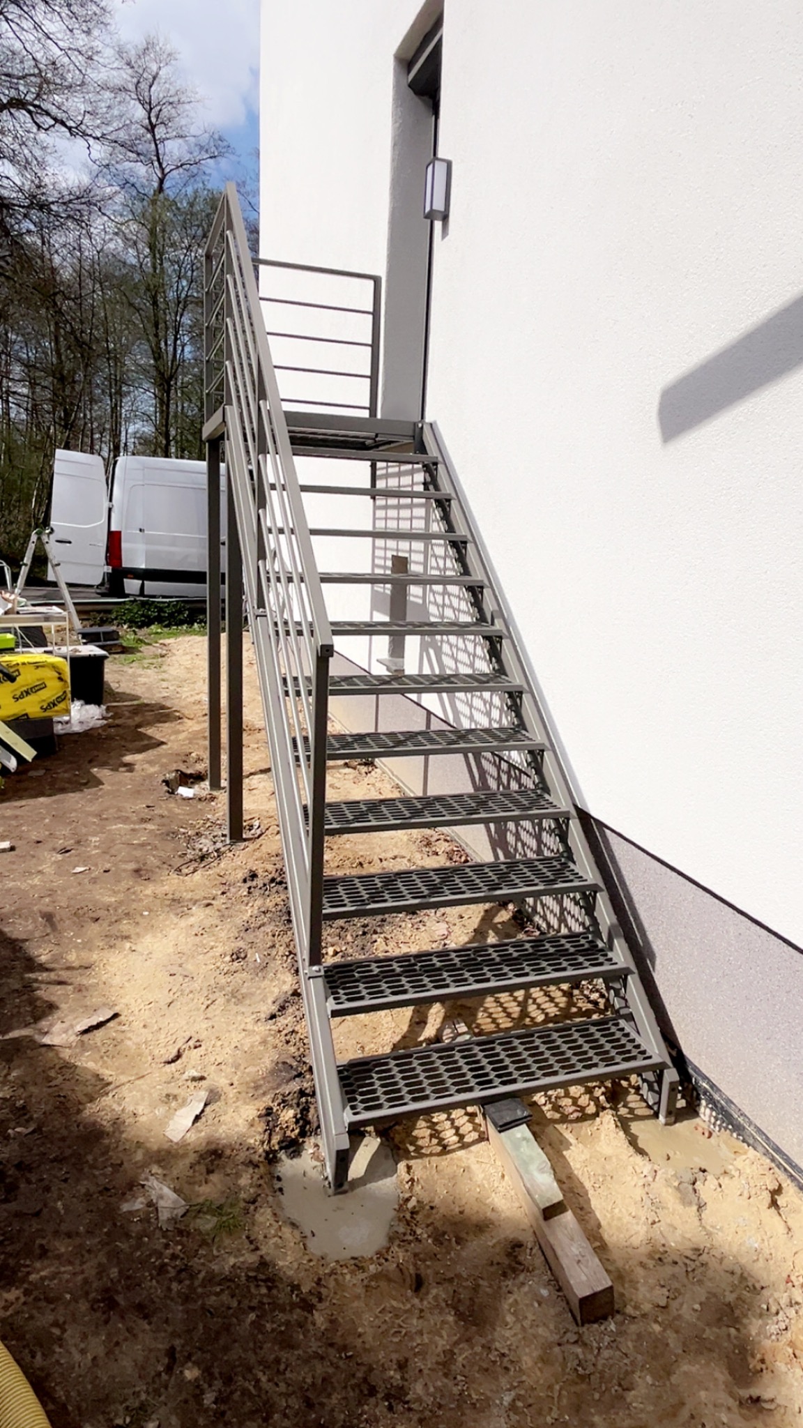 Zewnętrzne, metalowe schody z perforowanymi stopniami prowadzące do drzwi na piętrze budynku, widoczne ślady prac ziemnych wokół podstawy schodów.