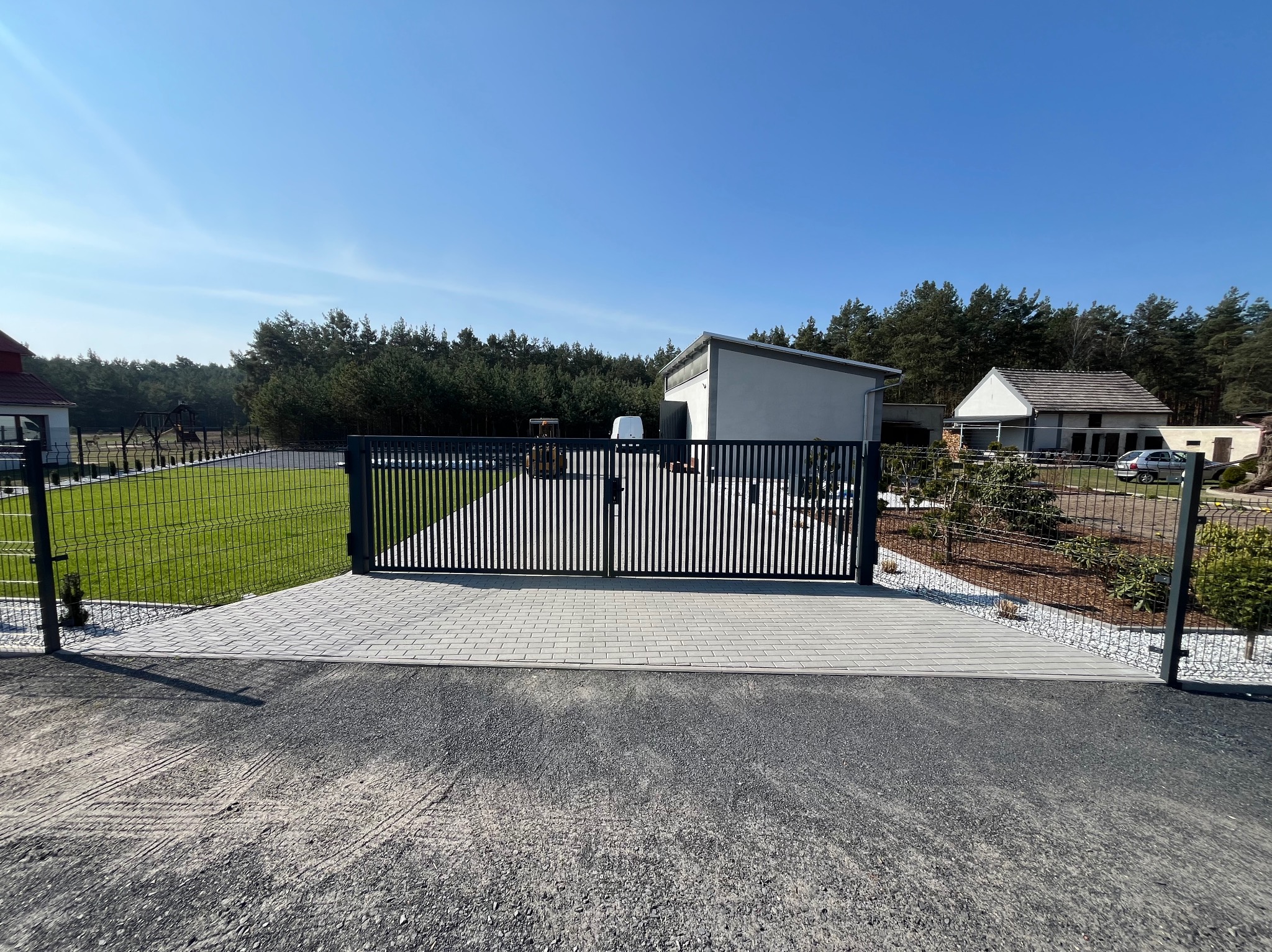 Nowoczesna brama wjazdowa z pionowymi profilami, widok z zewnątrz na posesję z kostką brukową, ogrodzeniem panelowym i budynkami w tle, słoneczny dzień.