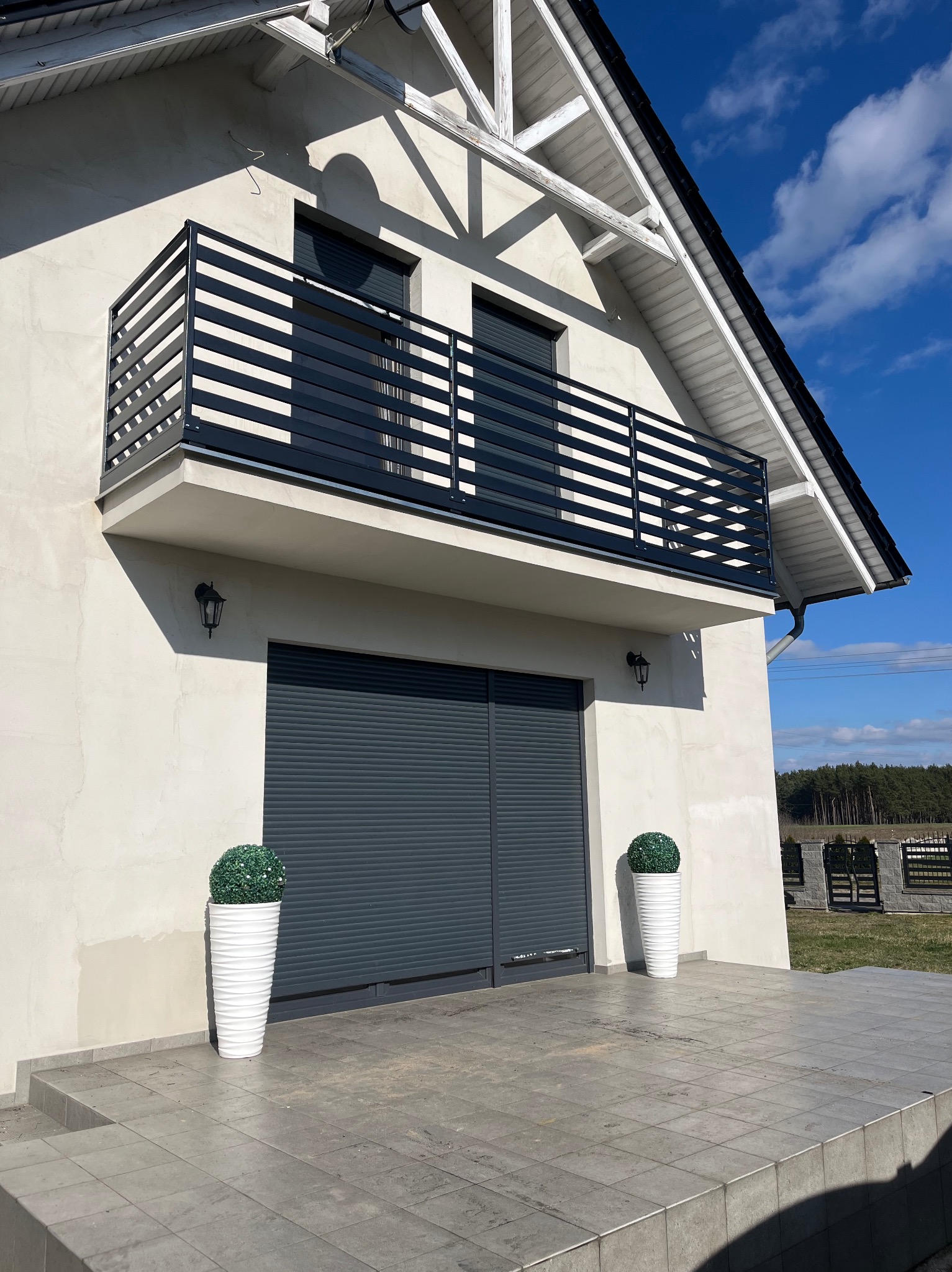 Nowoczesny dom z szarą, poziomą balustradą balkonową, szarą bramą garażową rolowaną i dwoma białymi donicami z kulistymi krzewami.