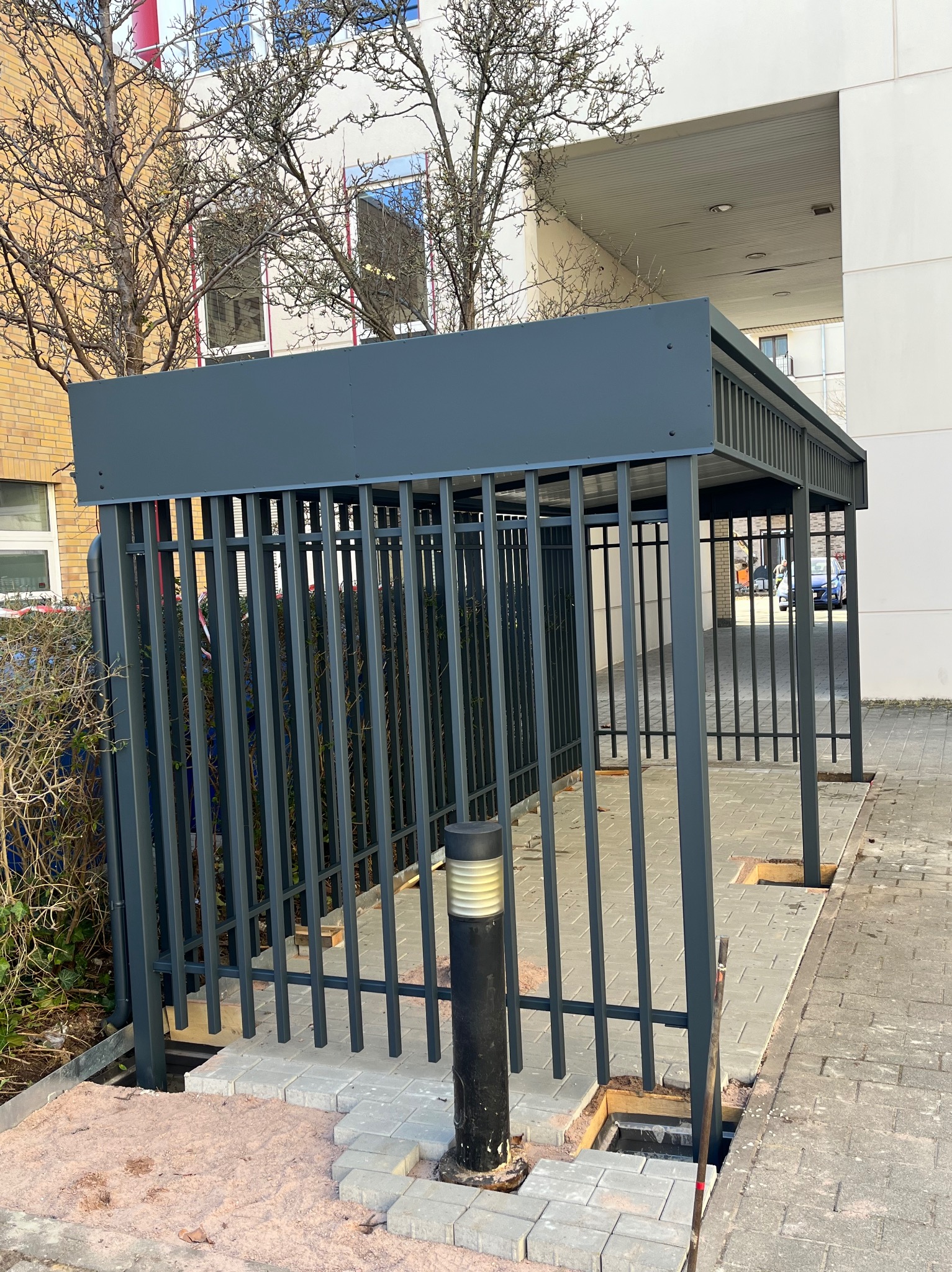 Szara, metalowa konstrukcja zadaszenia z pionowymi prętami, w trakcie montażu kostki brukowej wokół podstawy, widok na budynek w tle.