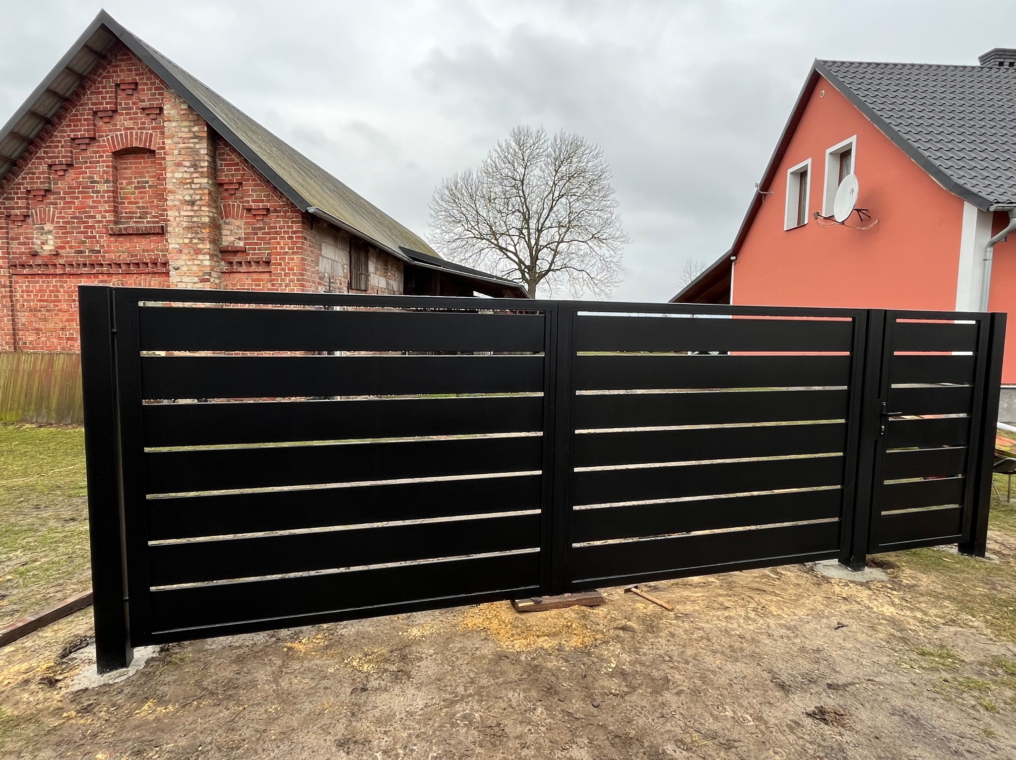 Nowoczesna brama wjazdowa przesuwna z czarnych, poziomych profili, na tle ceglanego budynku i domu z anteną satelitarną, w pochmurny dzień.