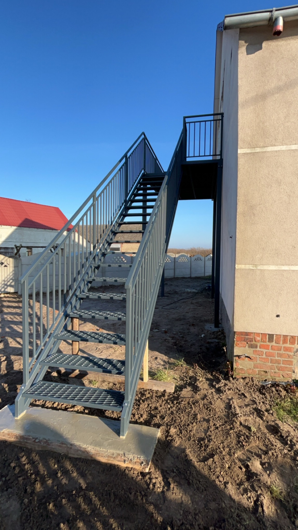 Zewnętrzne schody metalowe z ażurowymi stopniami, prowadzące na balkon, pomalowane na szaro, osadzone na betonowej podstawie, z widokiem na dom z czerwoną dachówką i błękitne niebo.