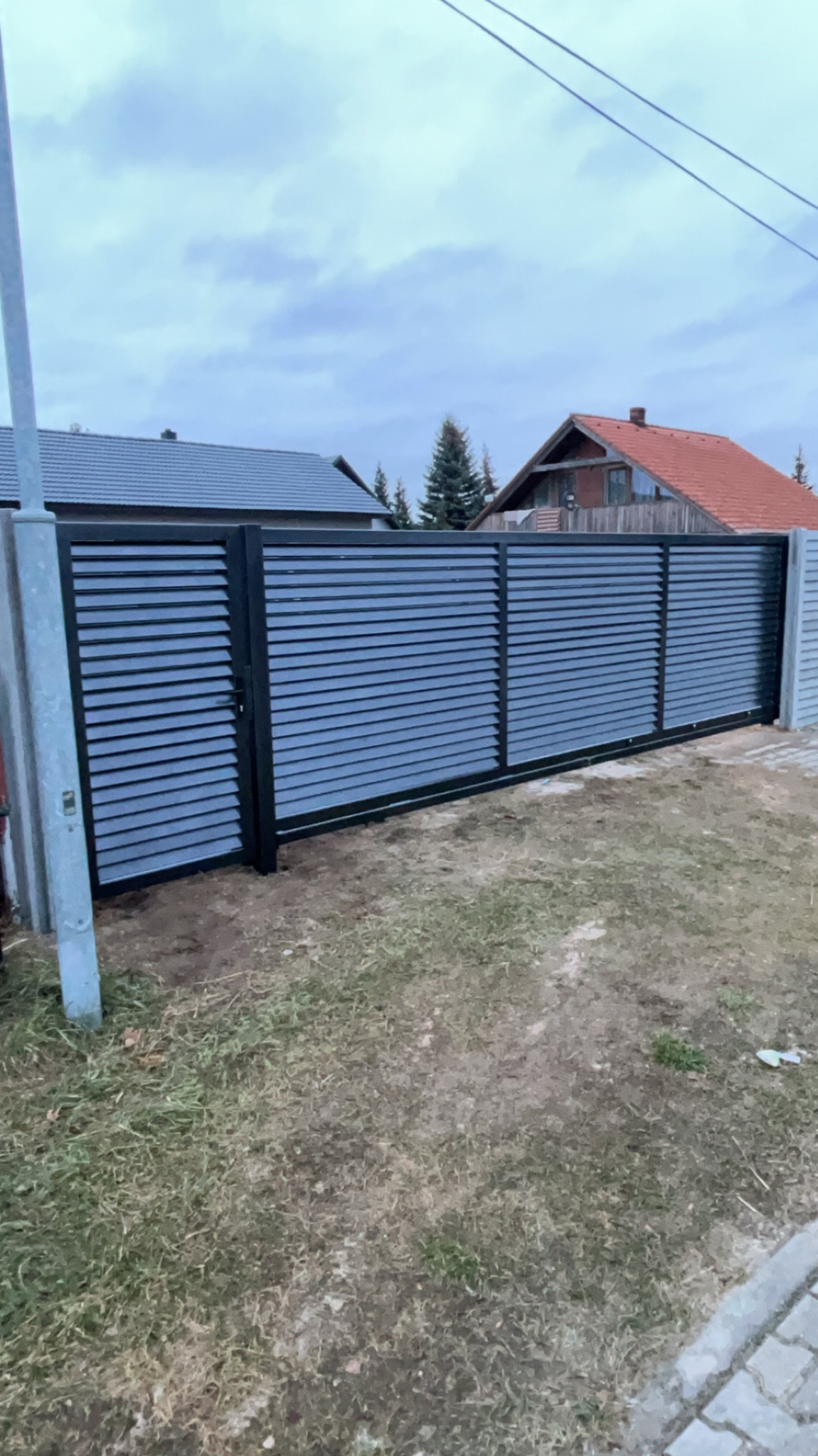 Przesuwna brama z poziomych, szarych profili, zamontowana między betonowymi słupkami, widok z zewnątrz na tle domów jednorodzinnych w pochmurny dzień.