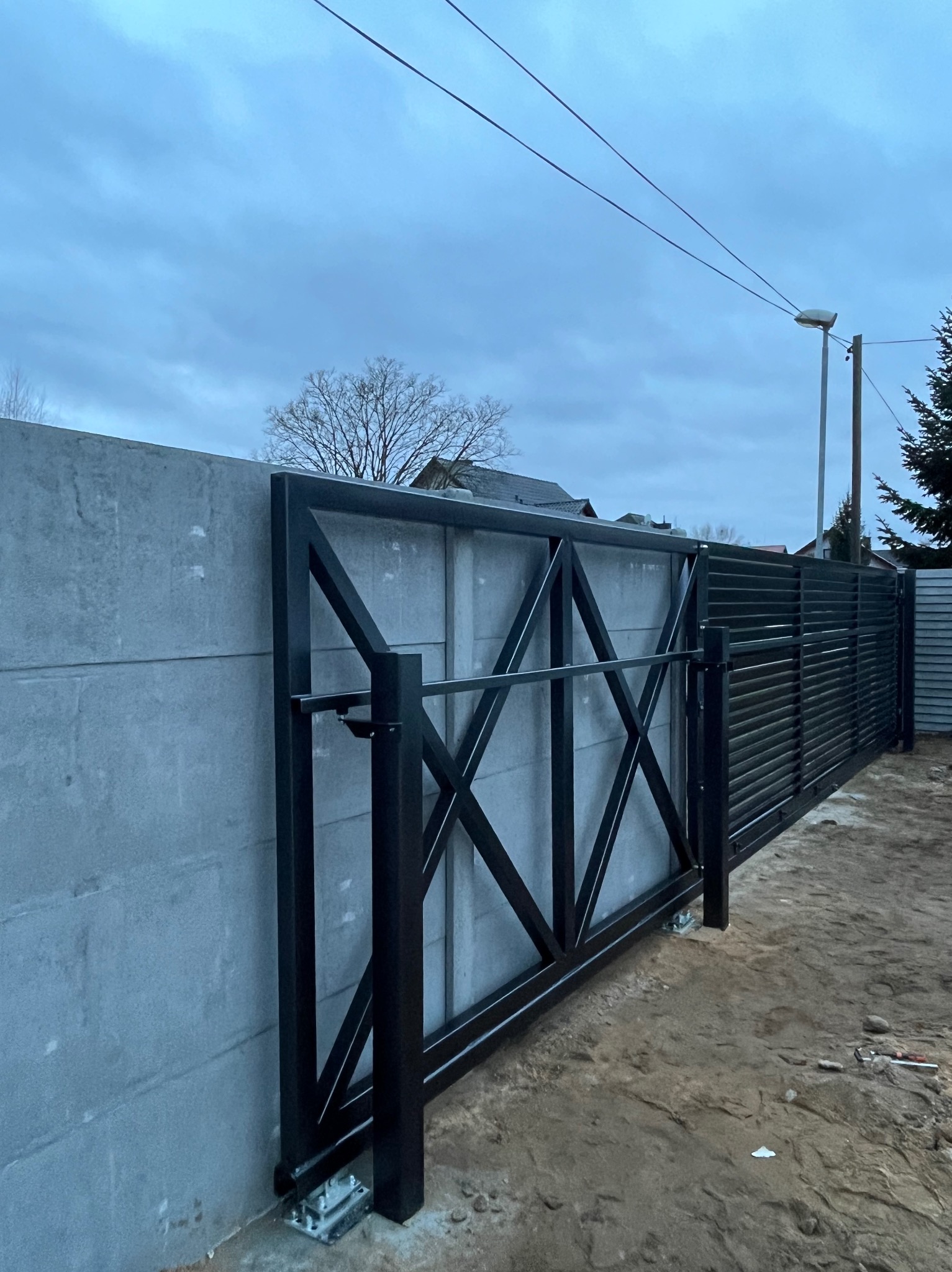 Przesuwna brama wjazdowa z czarną metalową konstrukcją w kształcie krzyża na betonowym murze, widok z boku na tle pochmurnego nieba.