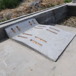 Betonowa konstrukcja z wbudowanymi otworami drenażowymi, prawdopodobnie część systemu odprowadzania wody, z ziemią i roślinnością w tle.