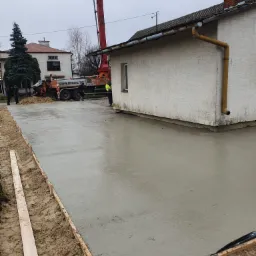 Świeżo wylany betonowy fundament pod budynek, z widocznym szalunkiem drewnianym i pompą do betonu w tle, na pochmurny dzień.
