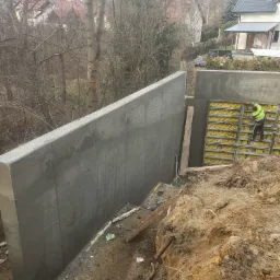 Betonowa ściana oporowa w trakcie budowy, widoczny pracownik w żółtej kamizelce montujący szalunki, z ziemią i drzewami w tle.