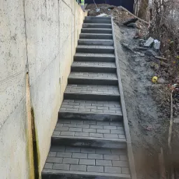 Schody z kostki brukowej prowadzące w górę, przylegające do betonowej ściany z widocznymi zaciekami, w tle pracownik z łopatą i ziemia.