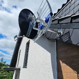 Antena satelitarna i kierunkowa zamontowana na dachu domu jednorodzinnego. Widoczne kable, mocowania i fragment elewacji budynku z oknami.