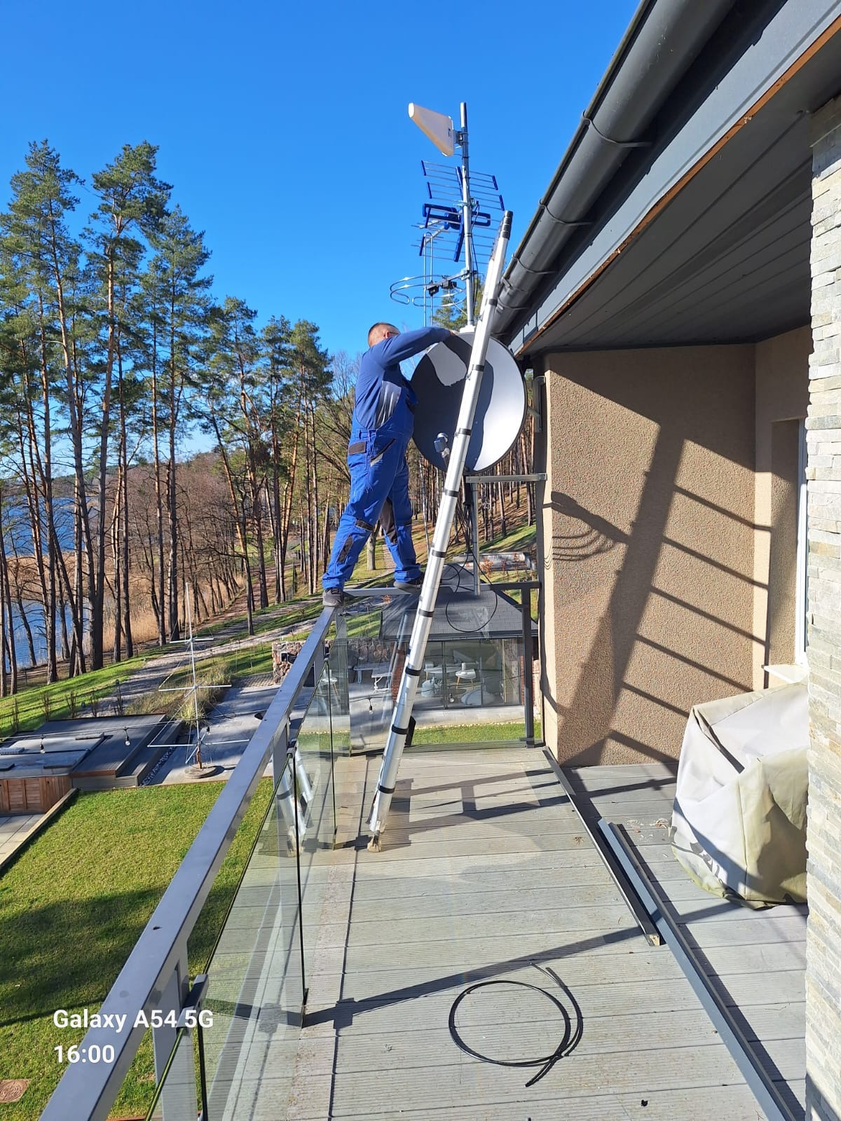Mężczyzna w niebieskim kombinezonie montuje antenę satelitarną na balkonie domu z widokiem na jezioro, używając drabiny.