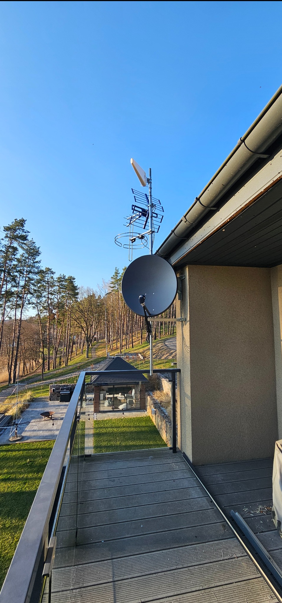 Antena satelitarna i naziemna zamontowana na balkonie nowoczesnego domu z widokiem na zalesione wzgórze i altanę. Błękitne niebo w tle.