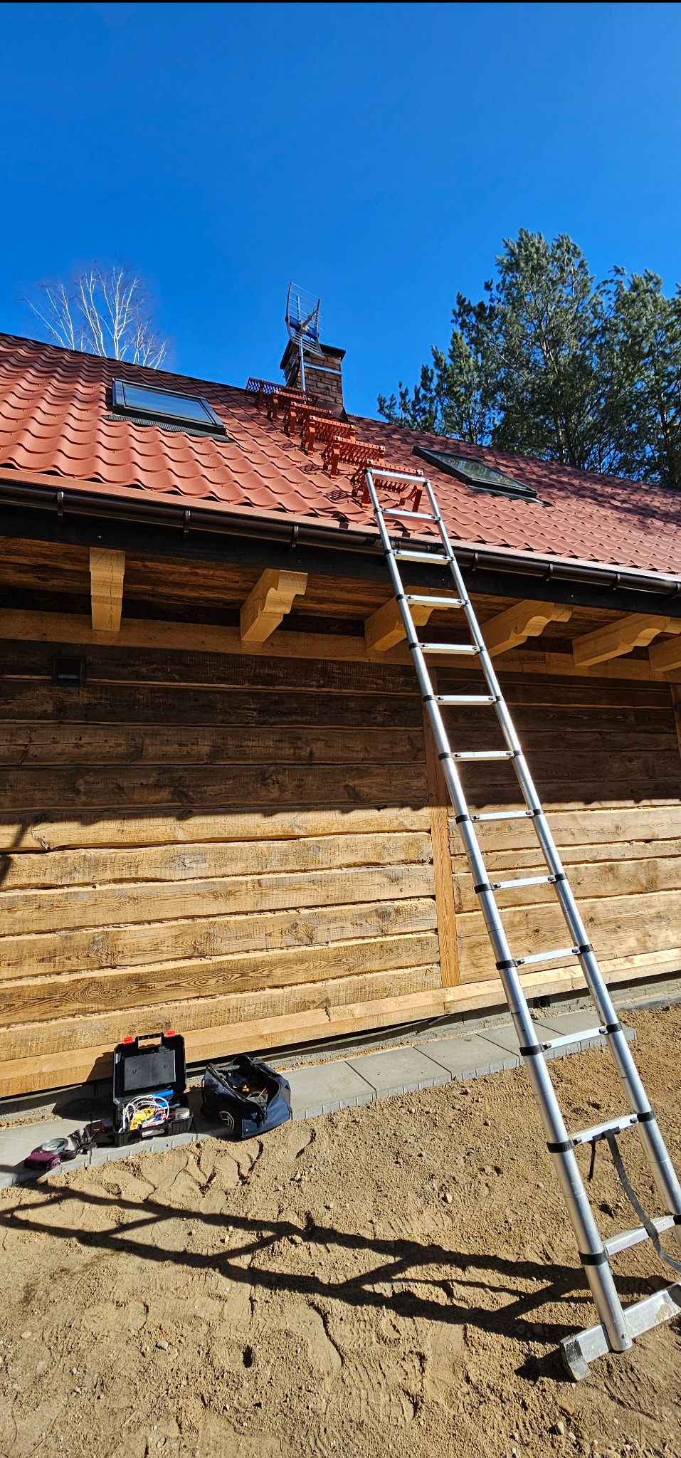 Montaż anteny na dachu domu w Jezioranach. Widoczna drabina oparta o drewnianą ścianę, narzędzia i antena na kominie. Czerwona dachówka i błękitne niebo.