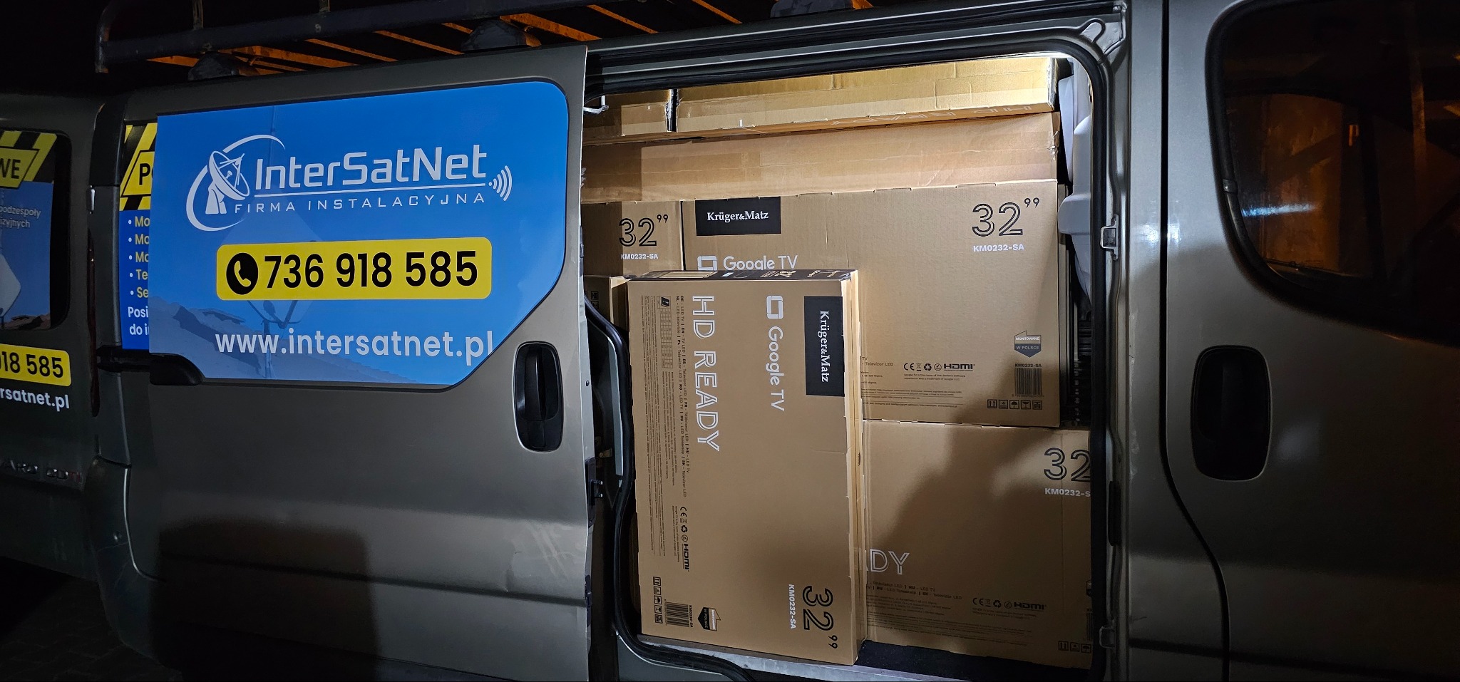 Furgonetka firmy instalacyjnej InterSatNet wypełniona kartonami telewizorów Kruger&Matz 32'. Widoczny numer telefonu i adres strony internetowej na boku pojazdu.