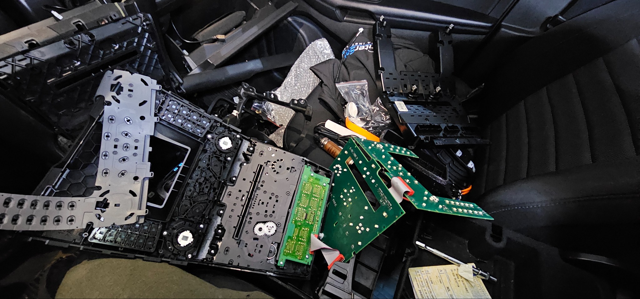 Rozłożony panel sterowania z elementami elektroniki, w tym zielone płytki drukowane i białe moduły, leżące na czarnym, teksturowanym tle, prawdopodobnie w samochodzie.