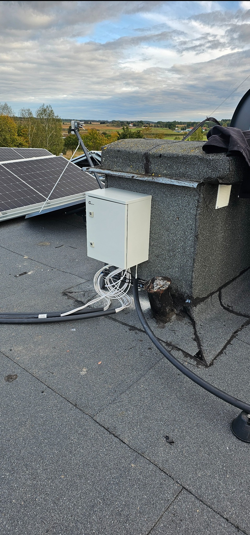 Biała skrzynka elektryczna zamontowana na dachu z licznymi kablami, panele słoneczne i antena satelitarna w tle, krajobraz za dachem.