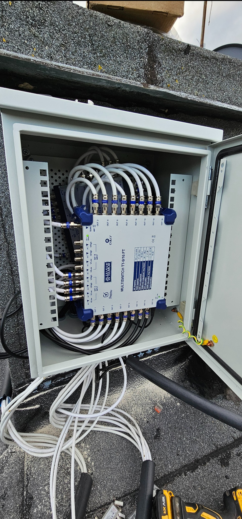 Rozdzielacz antenowy w szarej obudowie na dachu. Widoczne liczne kable koncentryczne, złącza i tabliczka znamionowa urządzenia Telkom-Telmor TT-9/16 FT.