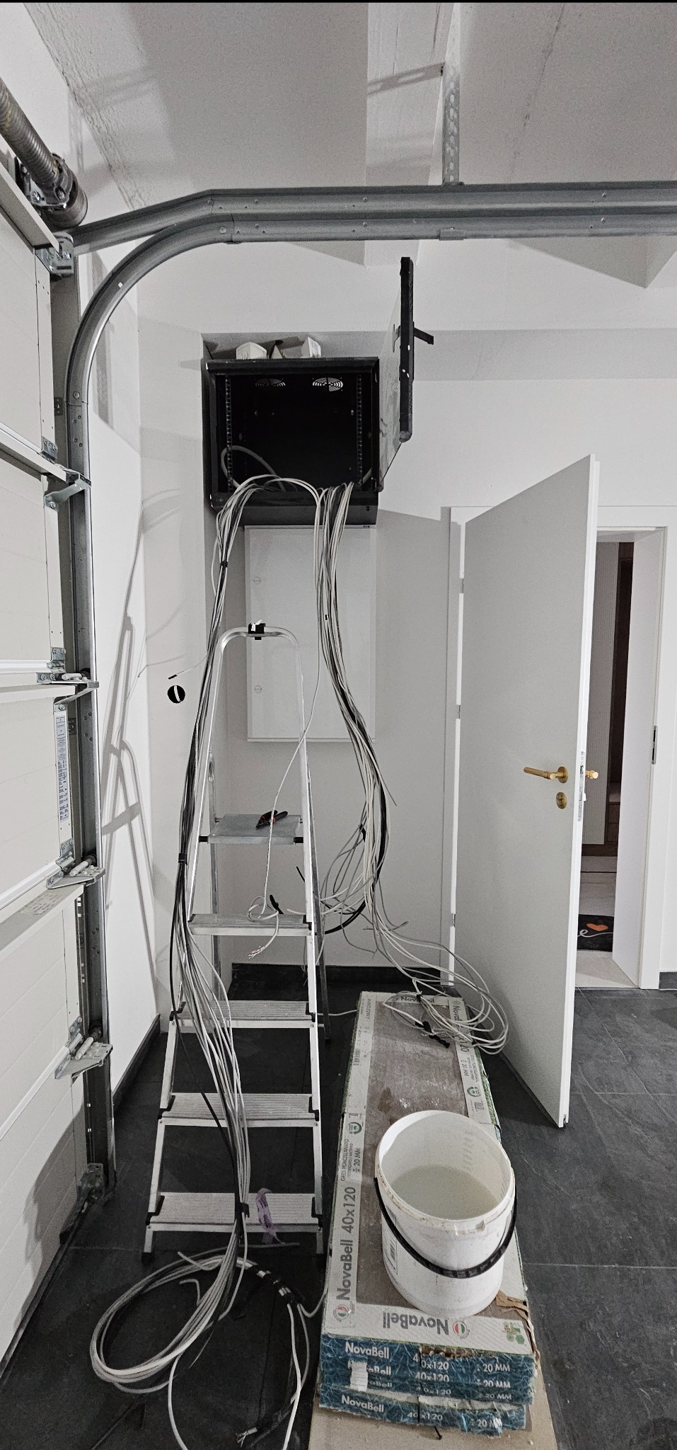 Montaż szafy serwerowej w garażu: widok na drabinkę, przewody i częściowo otwarte drzwi, ukazujący etap prac instalacyjnych w surowym wnętrzu.