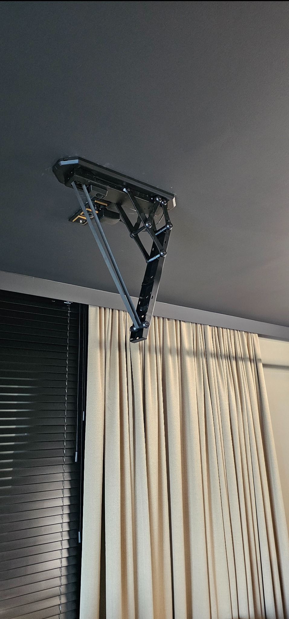 Czarny, metalowy uchwyt sufitowy na projektor, zamontowany w nowoczesnym wnętrzu z zasłonami i żaluzjami. Widok z dołu, ciemny sufit.