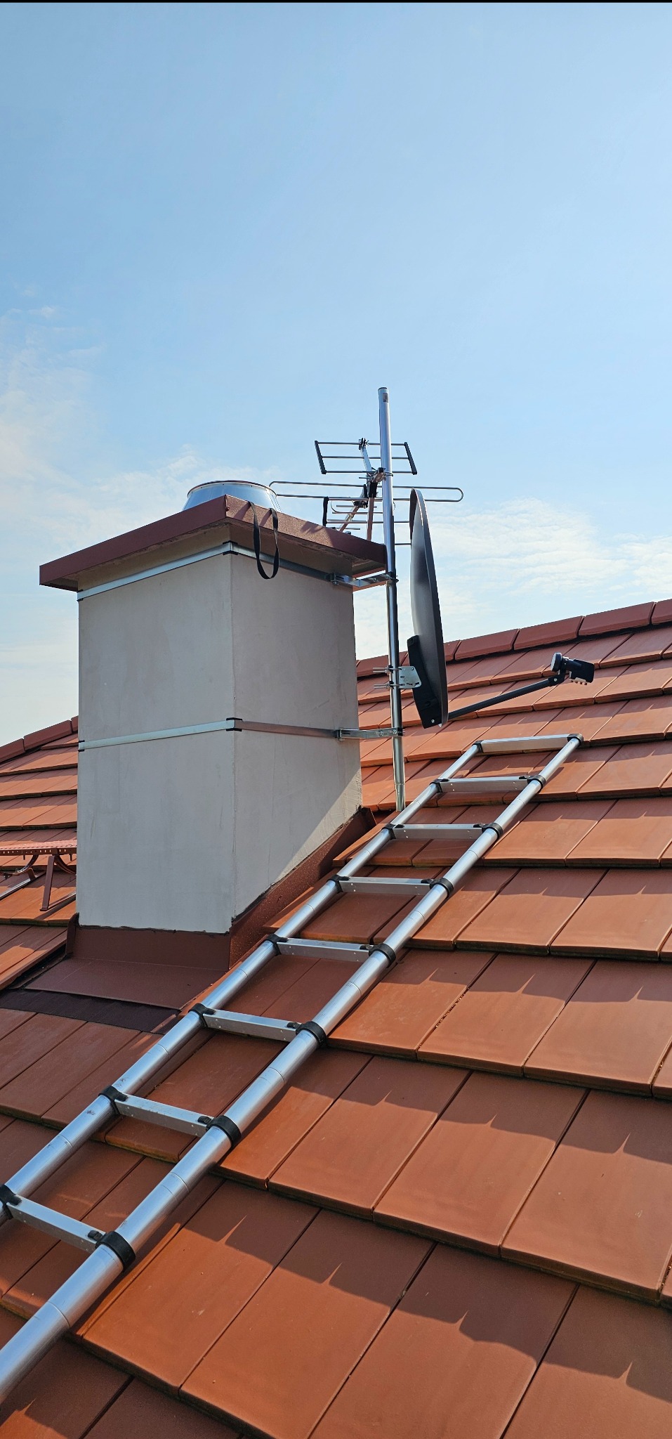 Antena satelitarna zamontowana na kominie, dostępna po rozłożonej aluminiowej drabinie na dachu z brązowej dachówki, na tle błękitnego nieba.