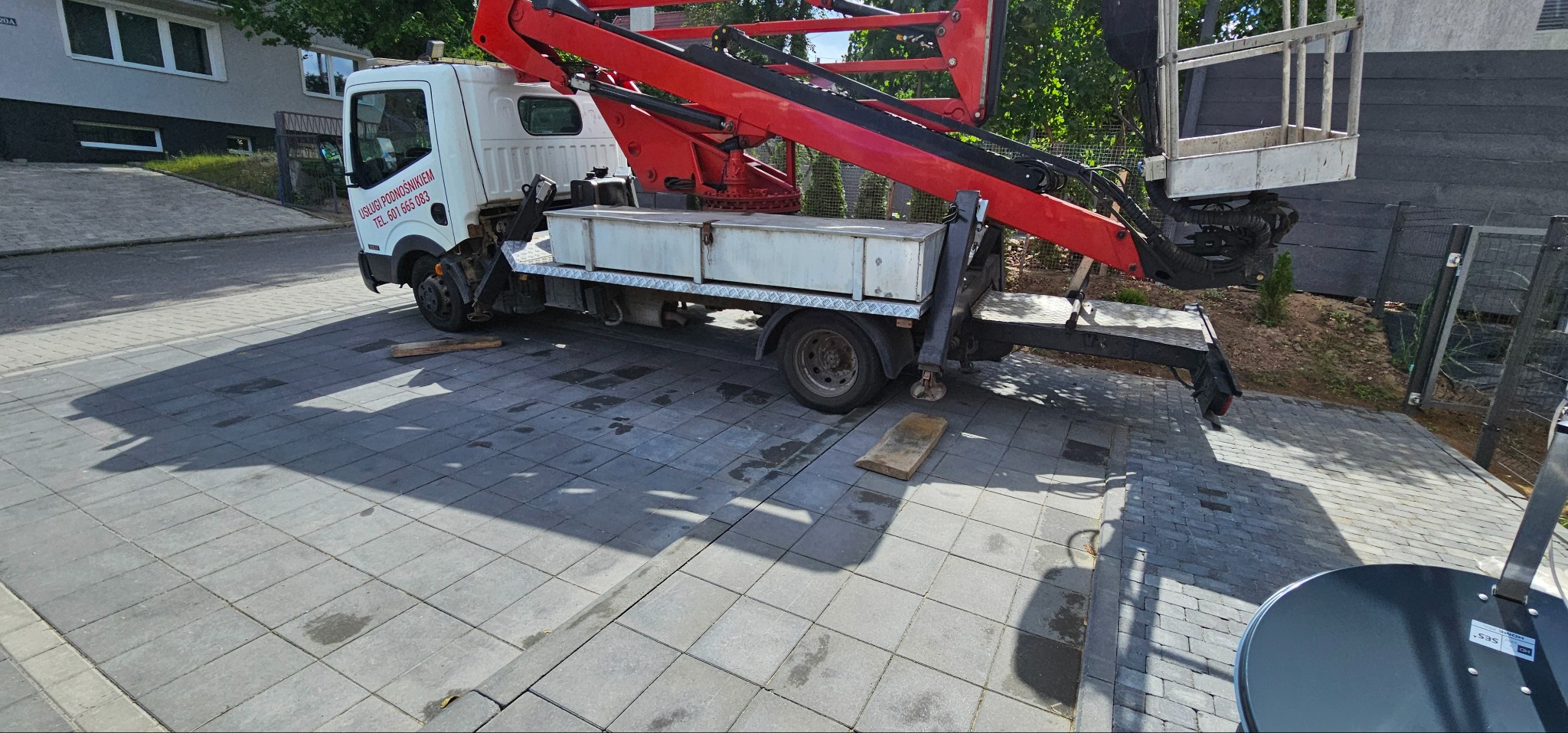 Samochód z podnośnikiem koszowym, zaparkowany na brukowanej powierzchni, z widocznymi elementami konstrukcji podnośnika i koszem. Usługi podnośnikiem, tel. 601 665 083.