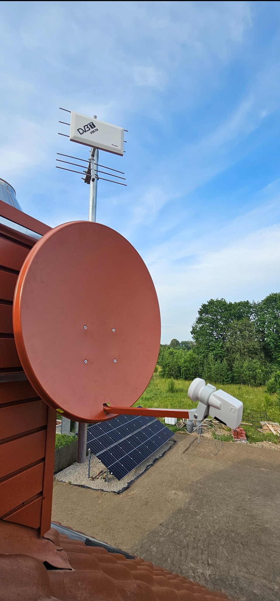 Antena satelitarna i naziemna DVB-T VISTA na dachu budynku z panelami słonecznymi w tle, z widokiem na zieloną okolicę i błękitne niebo.