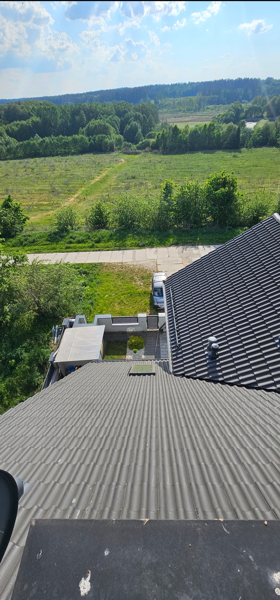 Widok z góry na szary dach z dachówki falistej, fragment ogrodu i krajobraz z lasem w tle, w słoneczny dzień.