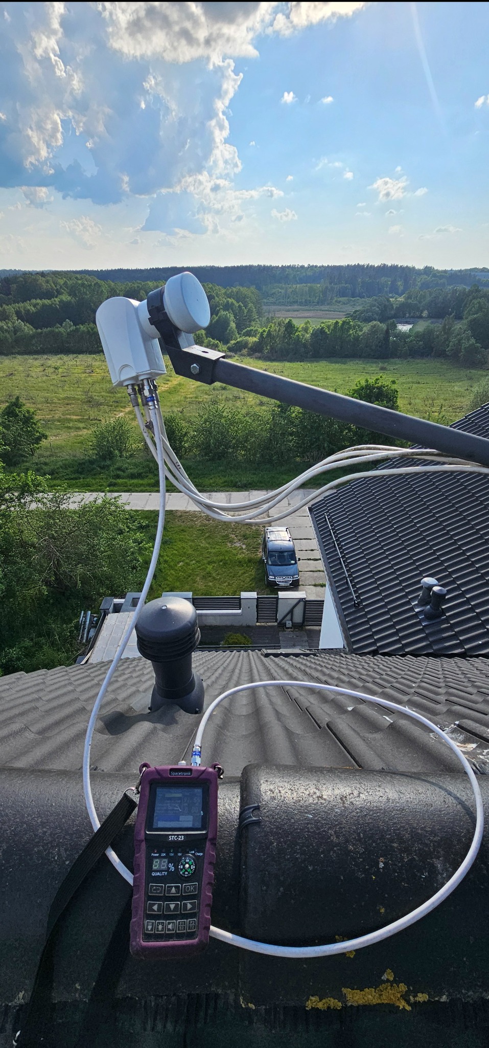 Antena satelitarna na dachu z analizatorem sygnału Spacetronik STC-23, widok na zielone tereny i błękitne niebo. Testowanie instalacji antenowej.