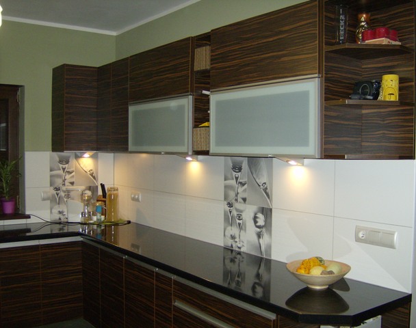 Nowoczesna kuchnia z ciemnobrązowymi szafkami o wzorze drewna zebry, czarnym blatem i białymi płytkami z motywem kieliszków, oświetlona halogenami podszafkowymi.