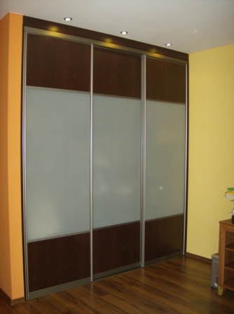 Trzydrzwiowa szafa przesuwna z brązowymi panelami i mlecznym szkłem, oświetlona punktowo, na tle pomarańczowej i żółtej ściany.