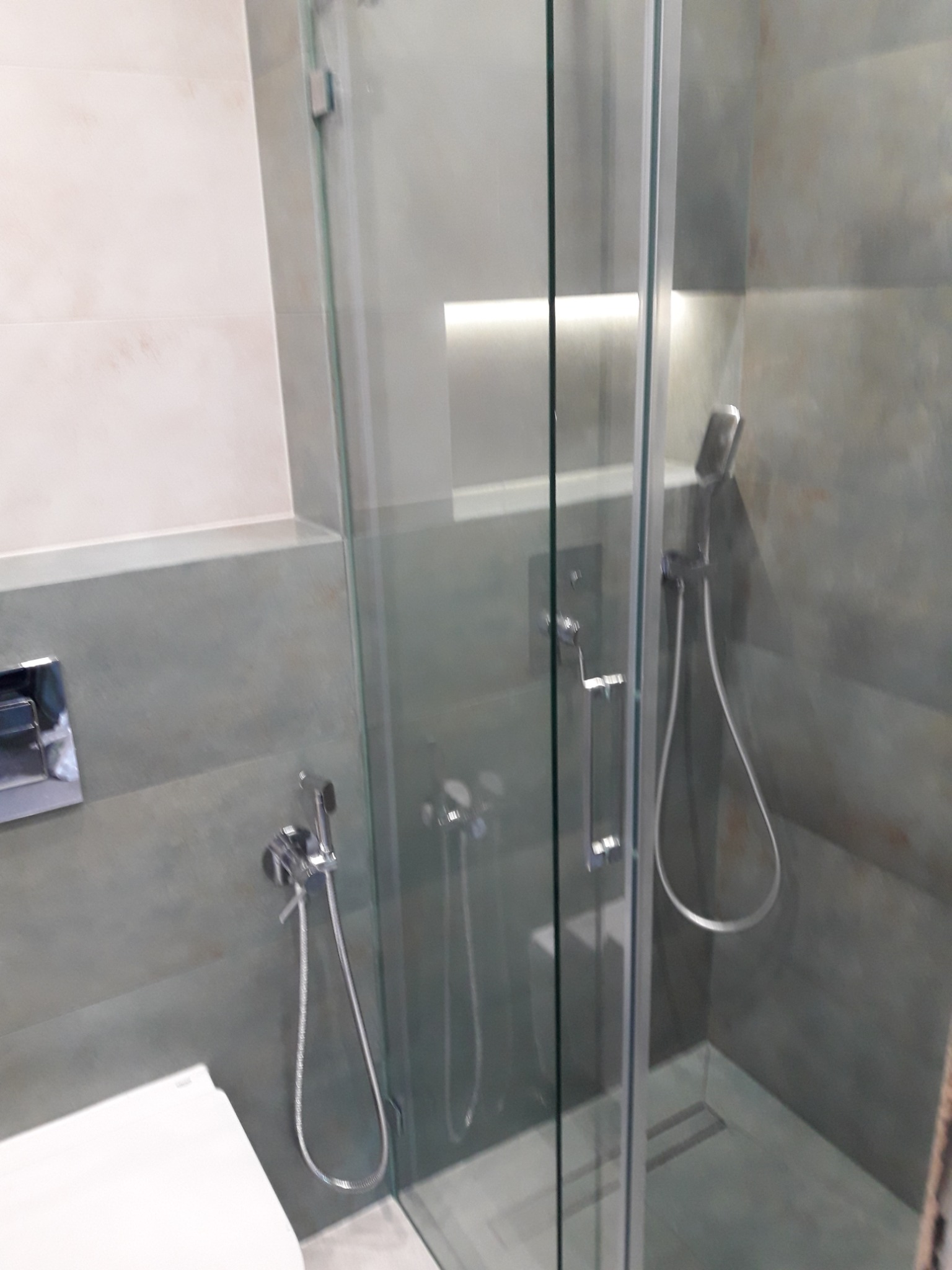 Szklana kabina prysznicowa w łazience z szarymi płytkami imitującymi beton, widoczny odpływ liniowy oraz bateria podtynkowa.