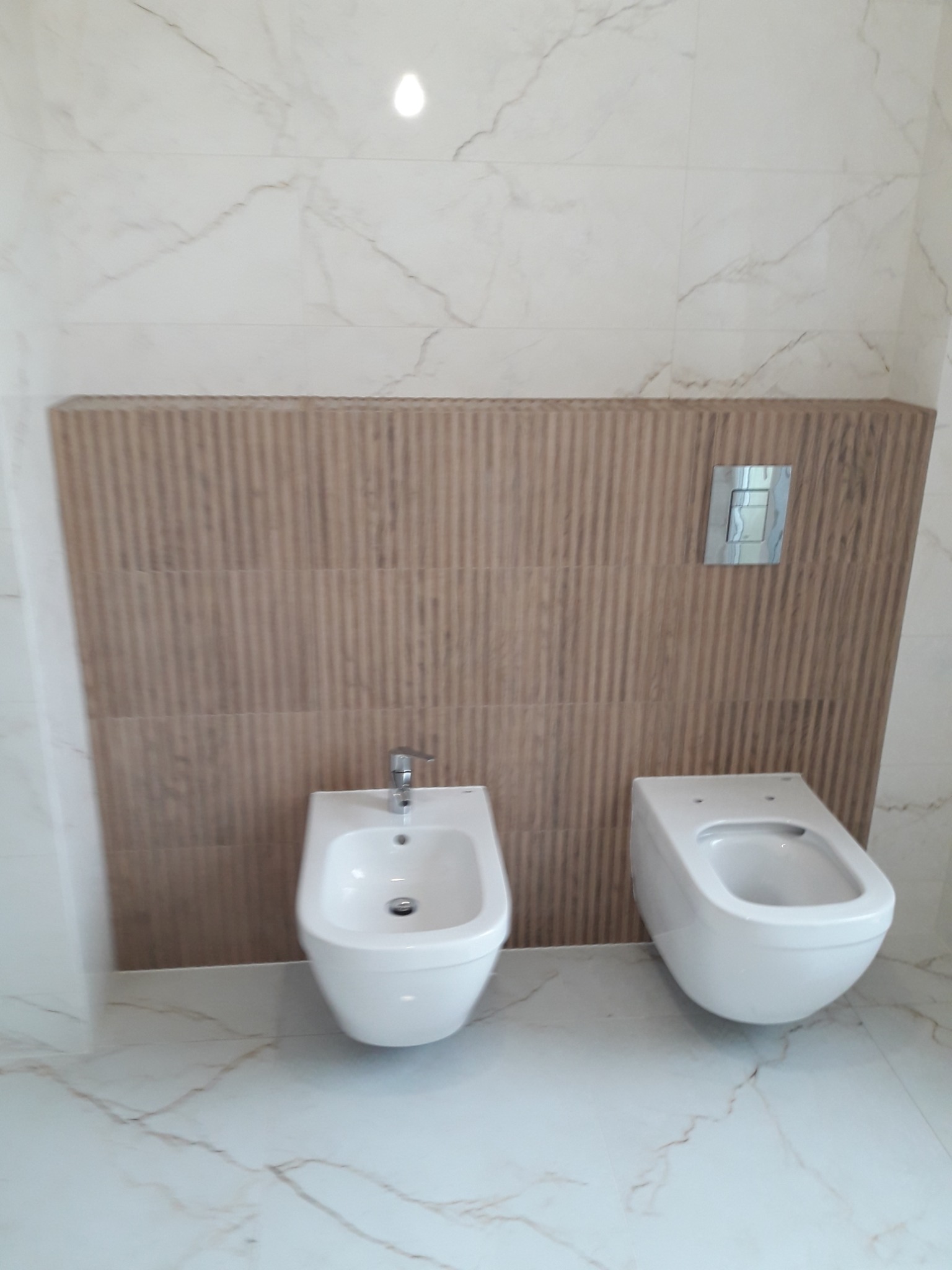 Bidet i toaleta podwieszana na tle ściany wyłożonej płytkami imitującymi drewniane panele, całość w łazience wykończonej marmurowymi płytkami podłogowymi i ściennymi.