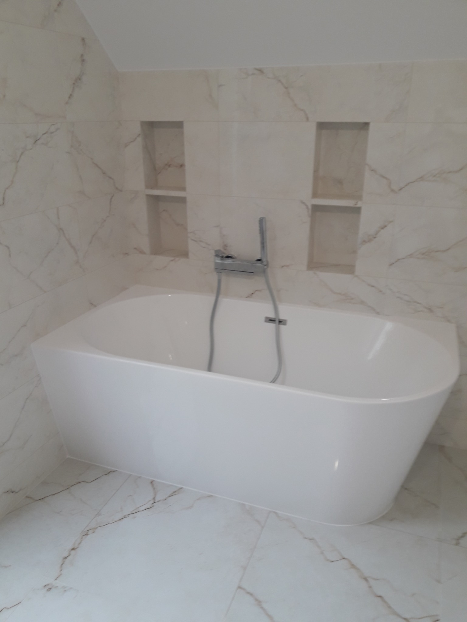 Nowoczesna, biała wanna wolnostojąca z chromowaną armaturą, otoczona marmurowymi płytkami z wnękami w ścianie, tworząc eleganckie wykończenie łazienki.