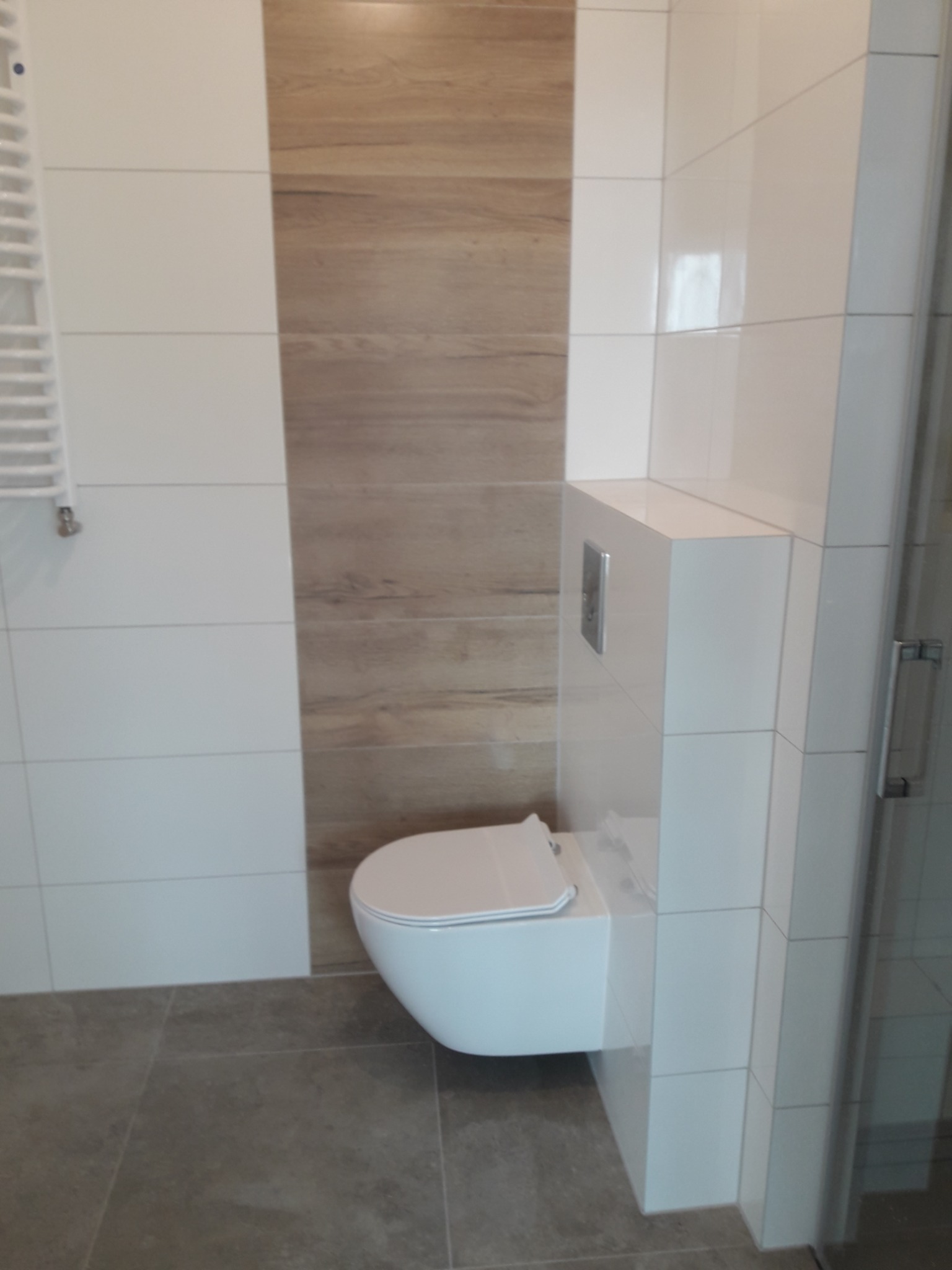 Nowoczesna łazienka z podwieszaną toaletą, białe i drewnopodobne płytki ceramiczne na ścianach, szare płytki podłogowe, grzejnik drabinkowy w tle.