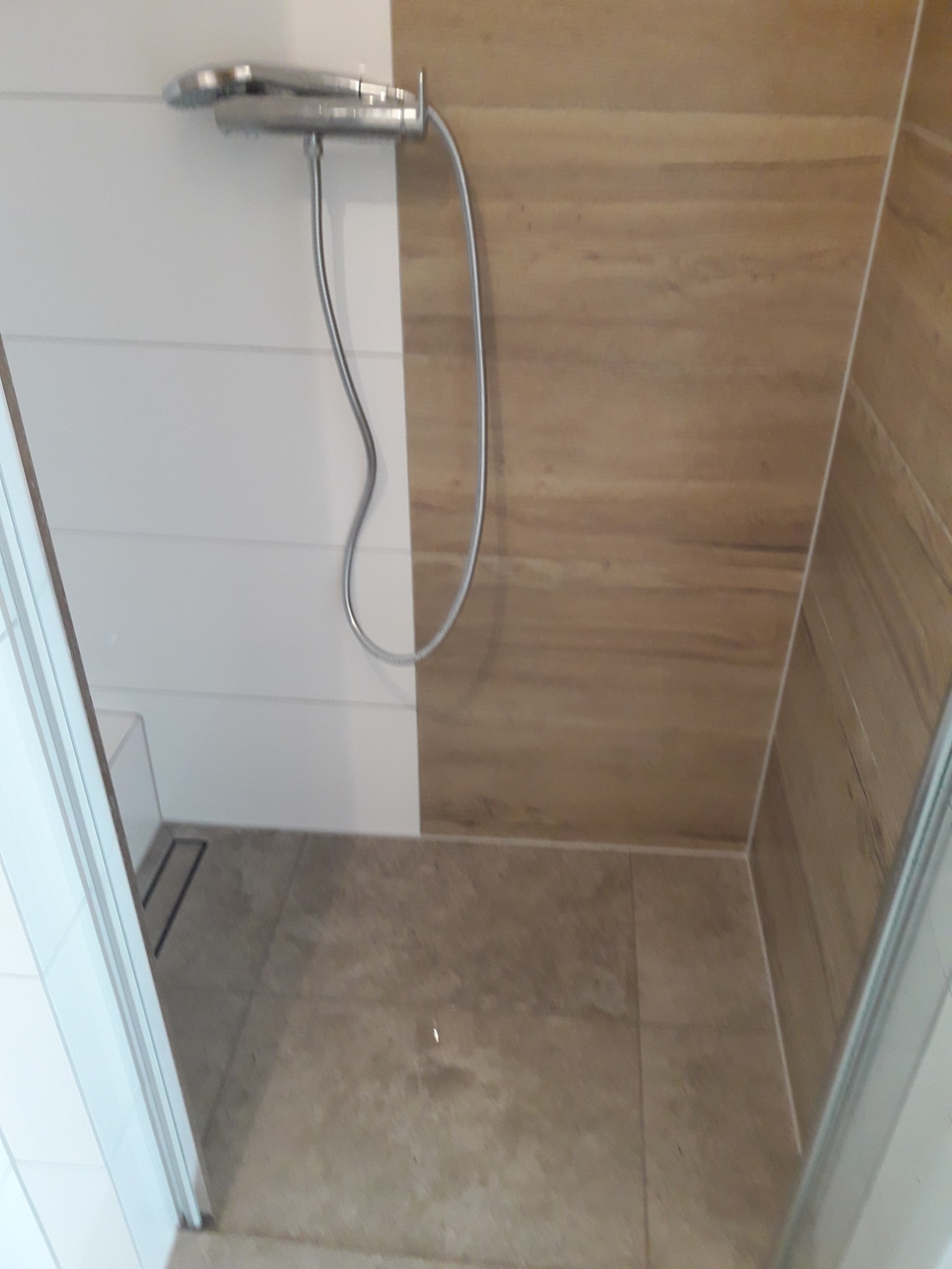 Kabiną prysznicowa z mieszanką płytek: białe poziome pasy i imitacja drewna, chromowana bateria natryskowa, odpływ liniowy w podłodze z beżowych płytek.