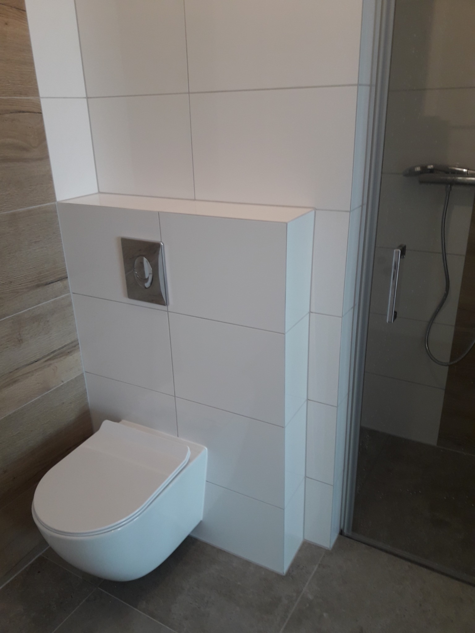 Nowoczesna, podwieszana toaleta z chromowanym przyciskiem spłukiwania, obudowana białymi płytkami, sąsiadująca z kabiną prysznicową. Widoczna imitacja drewnianej okładziny na ścianie po lewej stronie.