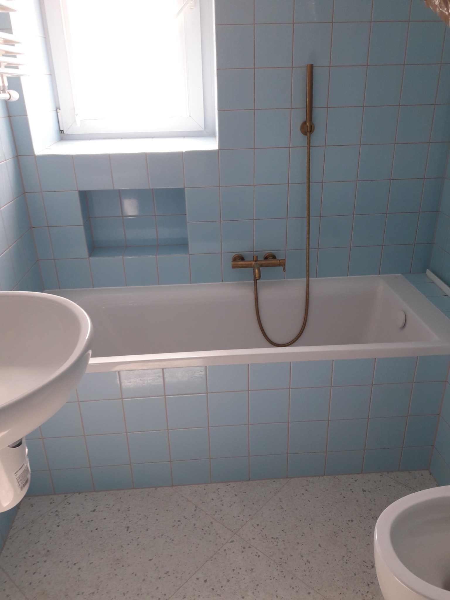 Łazienka z niebieskimi płytkami, białą wanną, umywalką i bidetem, z złotą armaturą i oświetleniem okiennym.