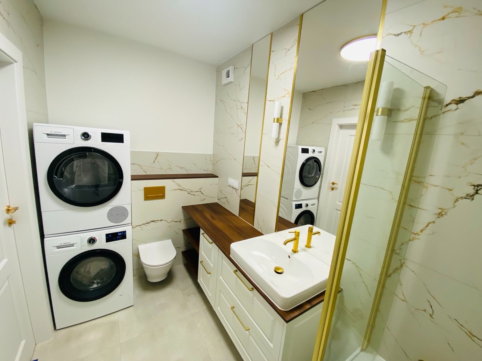 Nowoczesna łazienka z marmurowymi płytkami, złotymi akcentami, białą umywalką nablatową, szafką z szufladami, toaletą i pralko-suszarką Bosch ustawioną w pionie.