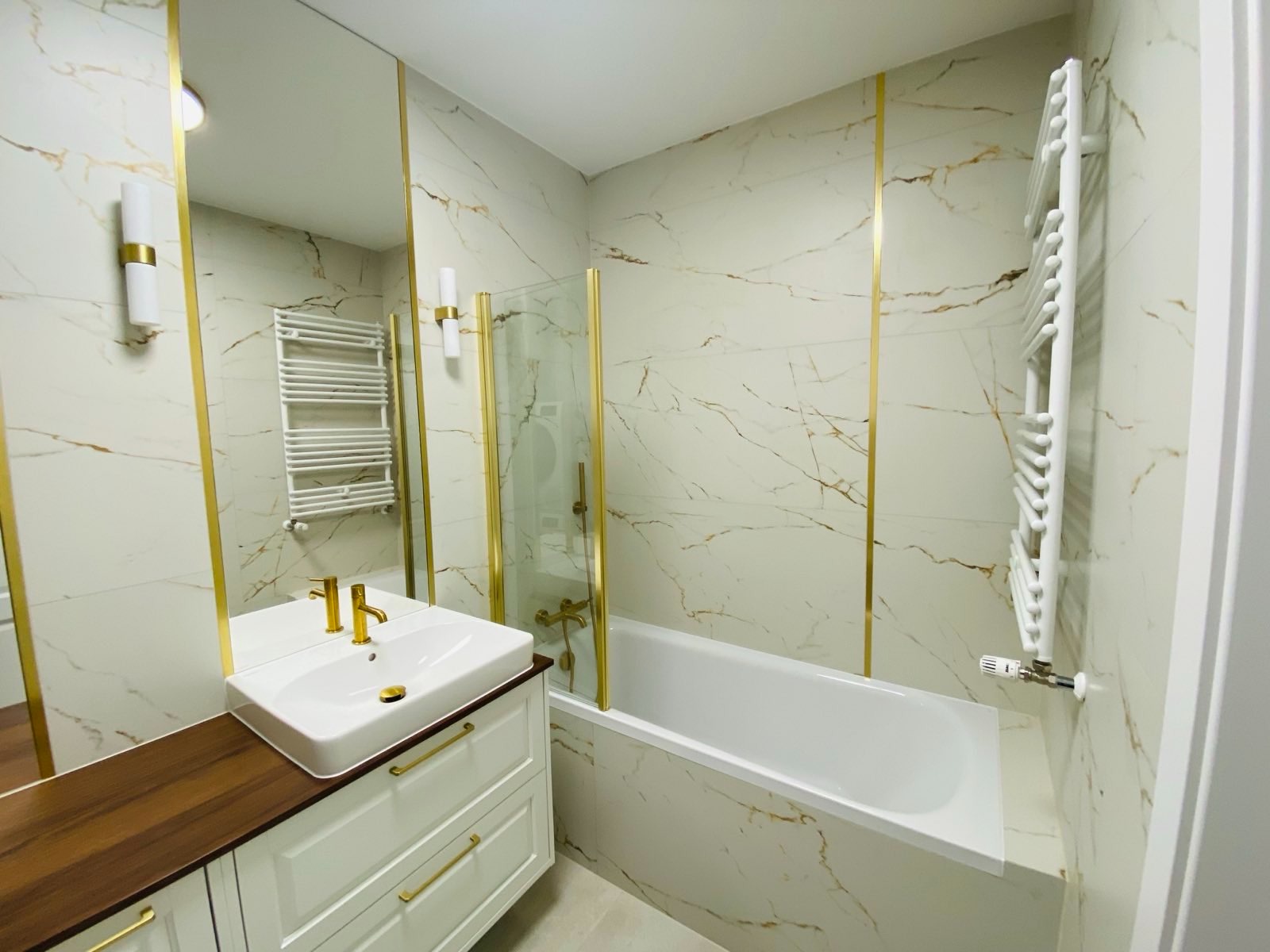 Nowoczesna łazienka z białymi płytkami imitującymi marmur, złotymi akcentami na armaturze, kabinie prysznicowej i listwach, białą szafką pod umywalką z drewnianym blatem i grzejnikiem drabinkowym