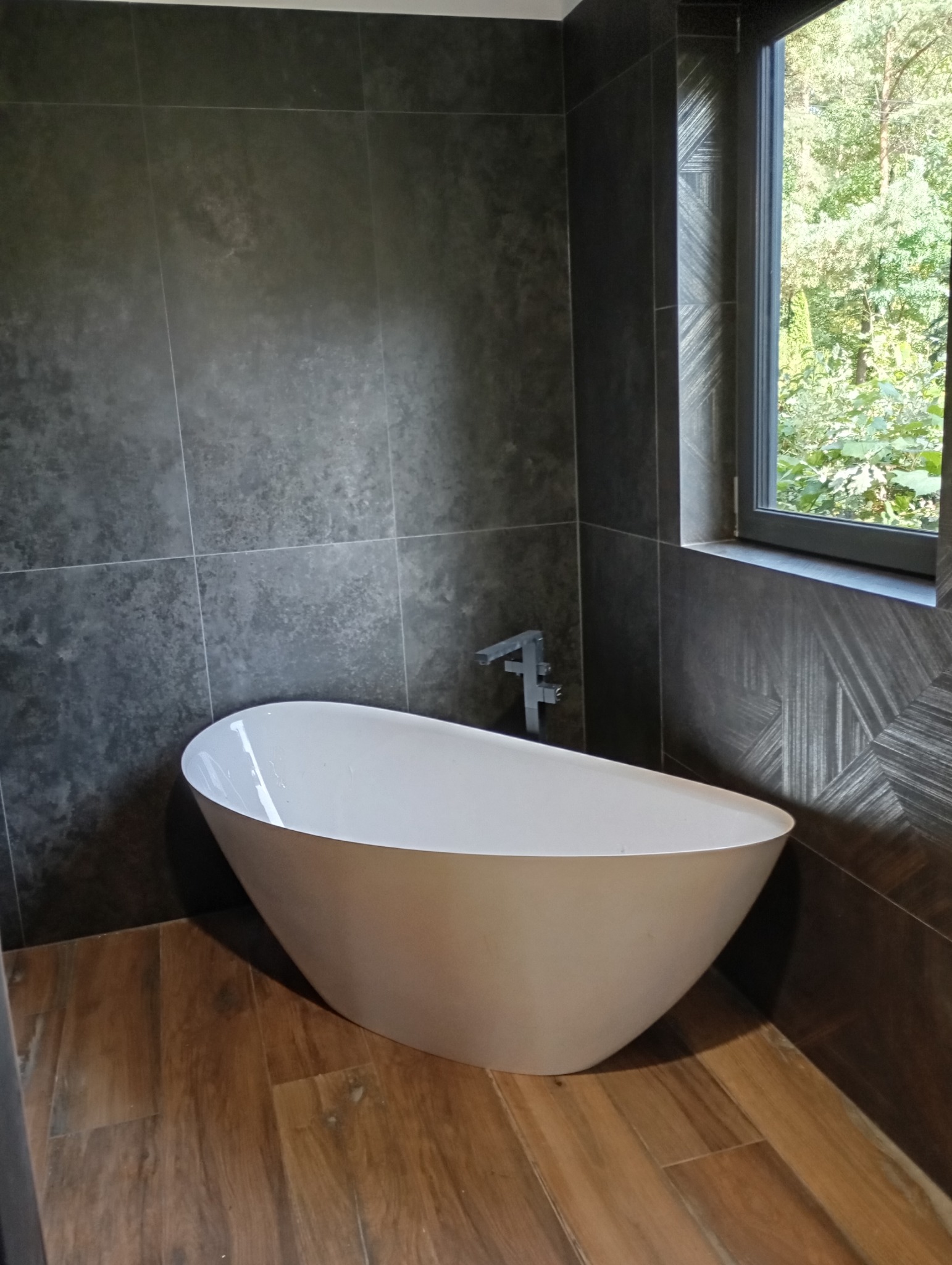 Nowoczesna łazienka z wolnostojącą białą wanną na drewnopodobnej podłodze. Ściany wyłożone ciemnymi, matowymi płytkami. Widok na zieleń przez okno.