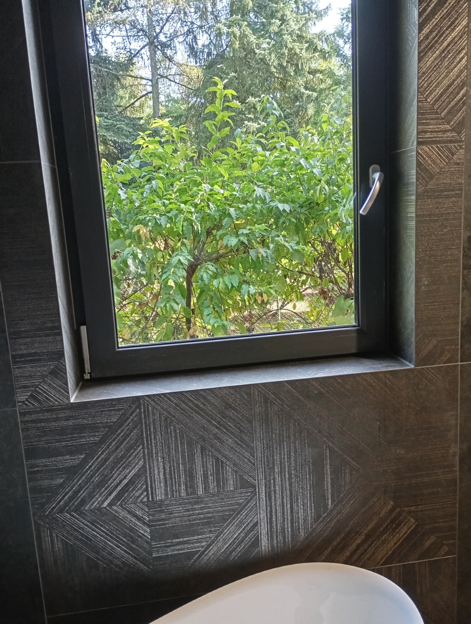 Łazienka z oknem: geometryczne, grafitowe płytki ścienne i widok na zielony ogród przez czarną ramę okienną. Część białej umywalki w dolnym rogu.