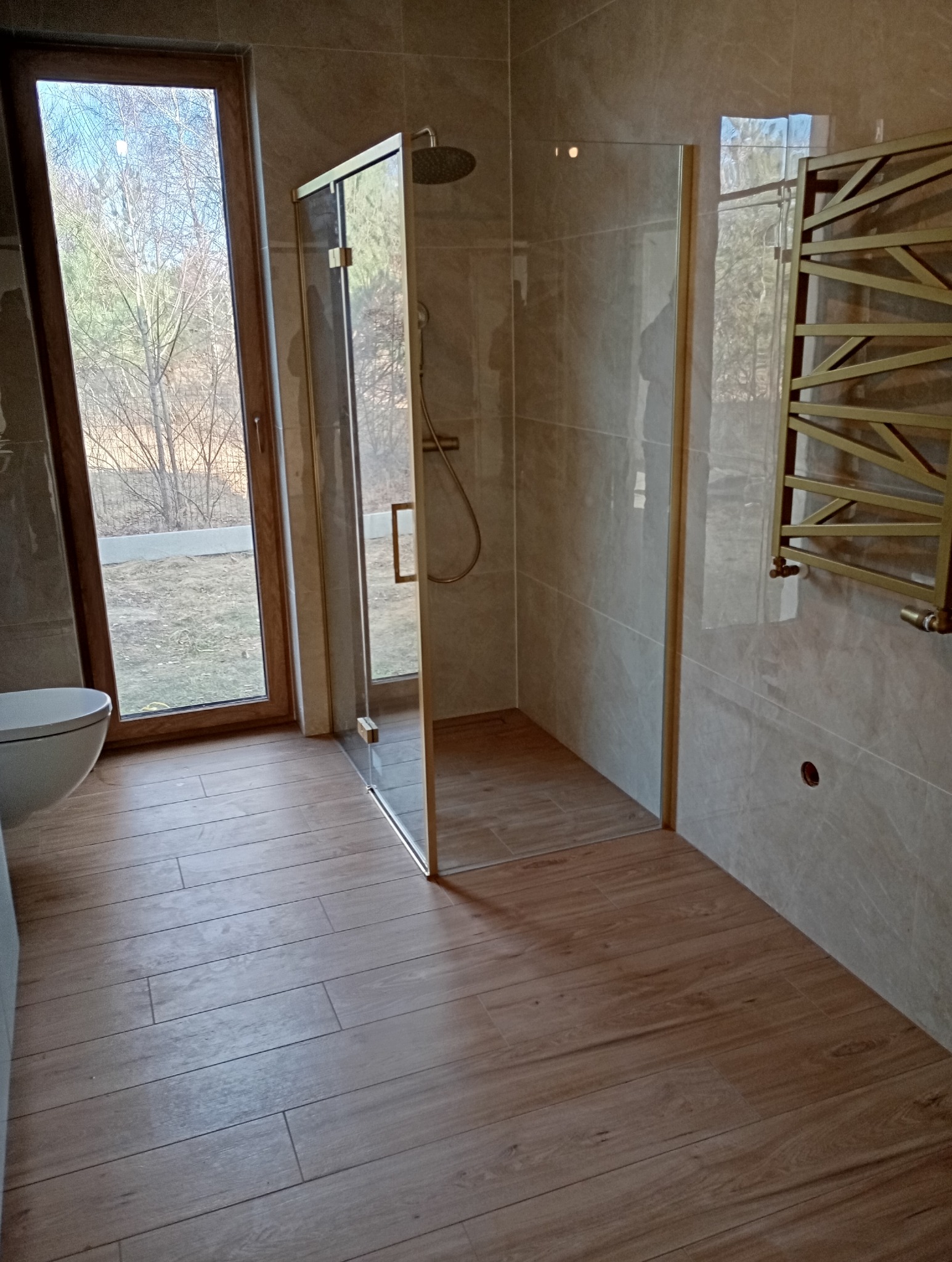 Nowoczesna łazienka z kabiną prysznicową typu walk-in, drewnopodobnymi płytkami na podłodze i grzejnikiem drabinkowym w kolorze złotym. Widok na okno.