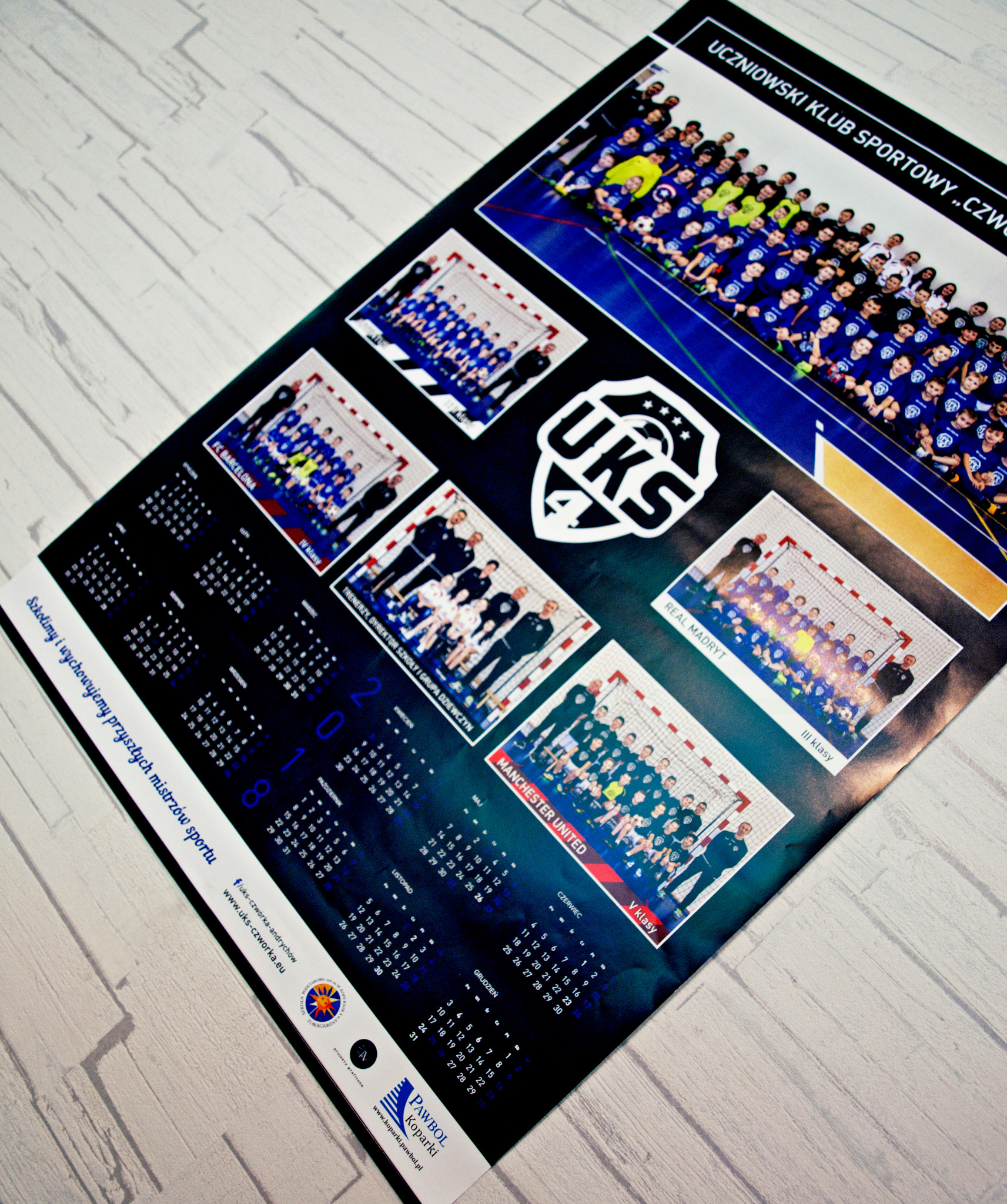 Kalendarz sportowy z fotografiami drużyn młodzieżowych i logo klubu sportowego UKS 4, leżący na jasnym tle imitującym drewno.