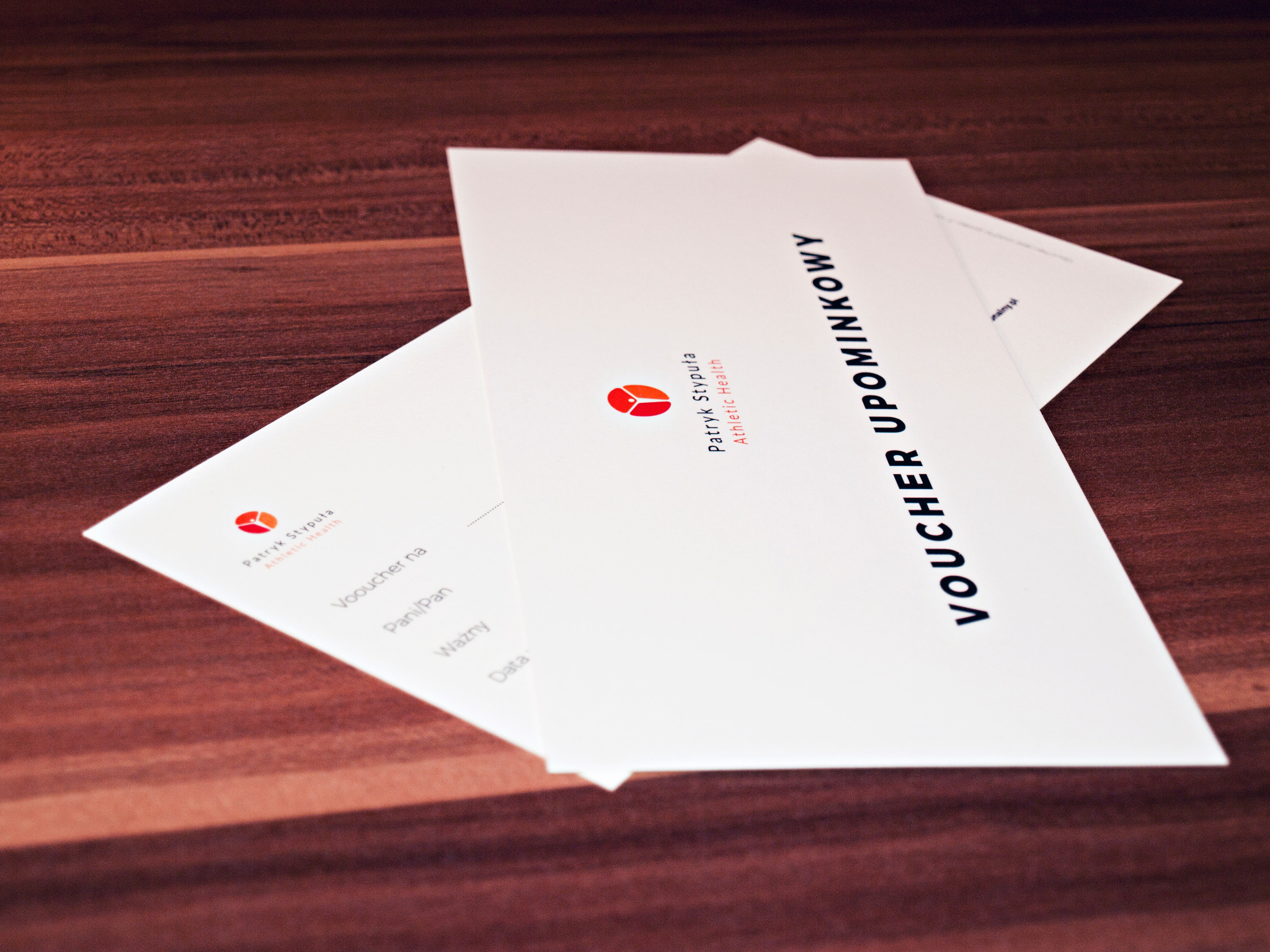 Dwa białe vouchery upominkowe z logo i danymi firmy Patryk Stypuła Athletic Health, leżące na drewnianym blacie.