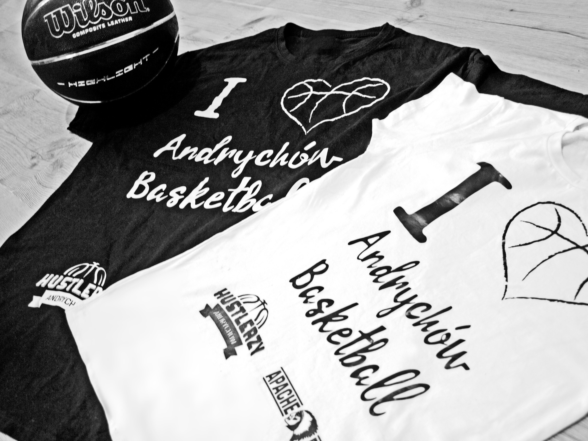 Dwie koszulki, czarna i biała, z nadrukiem 'I love Andrychów Basketball' oraz piłka do koszykówki Wilson na drewnianym tle.